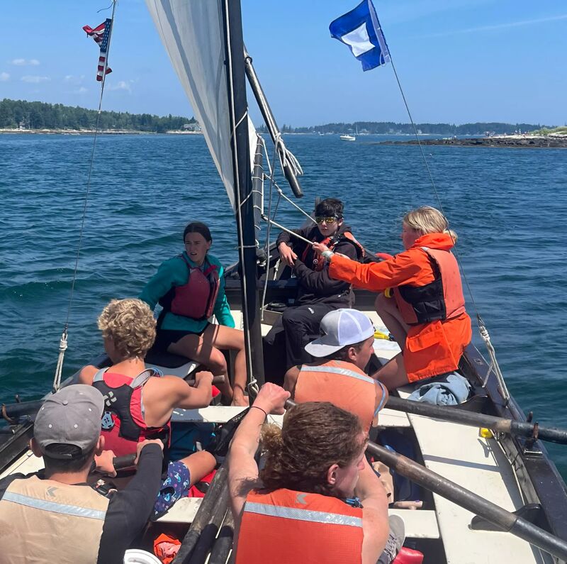 The image shows a group of people sailing on a small boat. They are on the water, with land visible in the background. The boat has a sail and several people are wearing life jackets. Some are seated while others are handling the boat's equipment. The sky is clear and the water is calm.